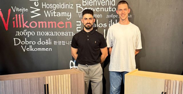 Two young men stand beside a wooden cabinet, smiling against a colorful multilingual welcome wall.