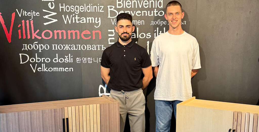 Two young men stand confidently beside a wooden cabinet against a welcoming multilingual wall. | © Name